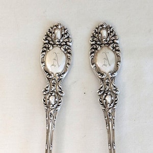 Puede incluir: Dos cucharas de servir antiguas de plata. Cada cuchara presenta un mango ovalado con un diseño floral ornamentado y una cartela central con la letra "A". Las cucharas tienen una superficie pulida y reflectante.