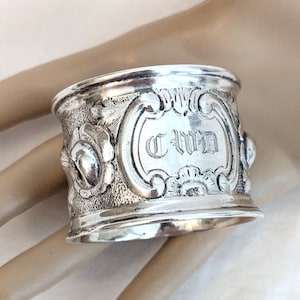 May include: A silver napkin ring with the name "Anna" engraved in an ornate font. The ring features floral and scrollwork designs. The napkin ring is held in a hand.
