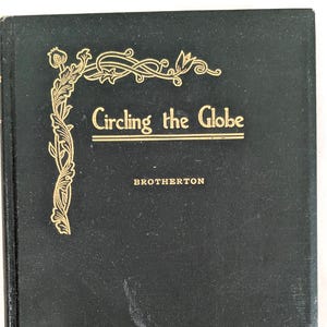 Könnte beinhalten: Ein dunkelgrünes Buch mit dem Titel "Circling the Globe" mit goldfarbenen Buchstaben und dekorativen Eckakzenten. Der Name des Autors, BROTHERTON, ist unter dem Titel aufgedruckt. Das Buch hat einen strukturierten Einband und weist Gebrauchsspuren auf.
