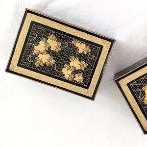 Könnte beinhalten: Zwei rechteckige schwarze Schachteln mit goldenen Rändern und Deckeln. Die Deckel weisen ein goldenes Traubenblatt- und Rankenmuster auf schwarzem Hintergrund auf. Die Schachteln sind vor einem weißen Hintergrund platziert.