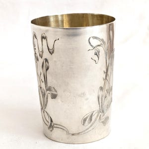 May include: A silver-coloured tumbler with an engraved floral design. The cup has a cylindrical shape with a slightly flared rim and a gold-coloured interior. The floral pattern is etched around the exterior of the cup.