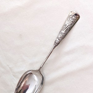 Puede incluir: Una cuchara de plata con un mango adornado con un diseño en relieve detallado de dos figuras. La cuchara tiene una forma ovalada clásica y un acabado pulido.