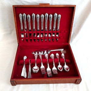 Puede incluir: Una caja de madera con un interior de terciopelo rojo, que contiene un juego de cubiertos de plata. Los cubiertos incluyen cuchillos, tenedores y cucharas, todos con mangos ornamentados.