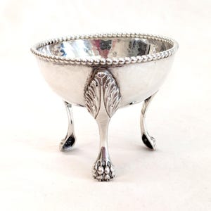May include: A small, silver-colored bowl with a hammered texture and a beaded rim. The bowl is supported by three ornate legs with a leaf design and claw feet. The bowl is likely made of metal and is designed for decorative use.
