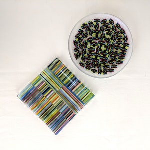 May include: Two glass art pieces on a white surface. One is a square dish with a woven pattern of colorful stripes. The other is a small, clear, round dish filled with small, colorful, rectangular glass pieces.