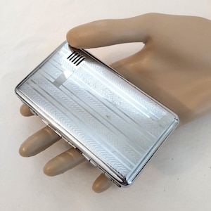 May include: A rectangular, silver-toned cigarette case with a textured, striped pattern. The case has a small vent on one end and a hinged opening. The case is held in a hand.