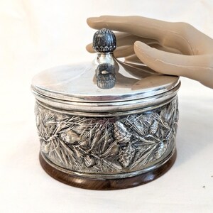 May include: A silver-colored round container with a lid and a wooden base. The container's side is decorated with a detailed relief of pine needles and pine cones. The lid has a small, decorative knob. The container is shown against a white background.