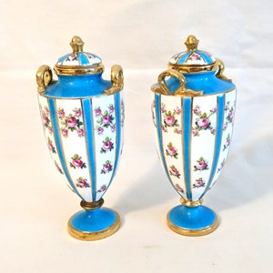 May include: A pair of blue and white porcelain vases with gold accents and floral designs. The vases have lids and are decorated with pink roses and gold stripes.