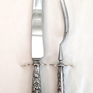 May include: A silver carving knife and fork set. The knife has a long, curved blade. Both pieces feature ornate floral designs on the handles and small, decorative guards near the blade and tines.