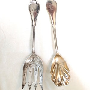 Puede incluir: Dos utensilios de servicio de plata, un tenedor y una cuchara, con mangos ornamentados y diseños grabados. El tenedor tiene tres dientes y la cuchara tiene un cuenco en forma de concha.