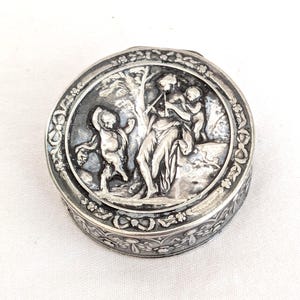 Puede incluir: Una caja de baratijas redonda de color plateado con una escena en relieve detallada de figuras tocando instrumentos musicales. La tapa presenta una imagen central rodeada por un borde floral. El lateral de la caja también está decorado con un motivo floral.