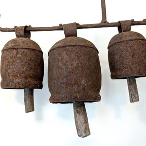 K&ouml;nnte beinhalten: Nahaufnahme eines Sets aus rustikalen Metallglocken. Die Glocken sind in verwittertem Braun gehalten und haben Holzkl&ouml;ppel. Sie sind an einer passenden Metallstange aufgeh&auml;ngt. Die Glocken haben ein Vintage- und antikes Aussehen.
