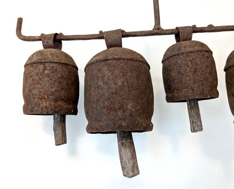 K&ouml;nnte beinhalten: Nahaufnahme eines Sets aus rustikalen Metallglocken. Die Glocken sind in verwittertem Braun gehalten und haben Holzkl&ouml;ppel. Sie sind an einer passenden Metallstange aufgeh&auml;ngt. Die Glocken haben ein Vintage- und antikes Aussehen.