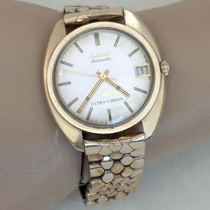 May include: A gold-tone Longines Ultra-Chron automatic wristwatch with a white dial and gold-tone hands. The watch has a date window at the 3 o'clock position and a gold-tone bracelet.