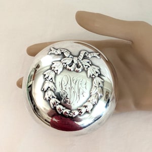 May include: A round, silver compact with a decorative wreath and ribbon design. The center features engraved initials. The compact is highly reflective, with a polished surface. A classic, elegant accessory.