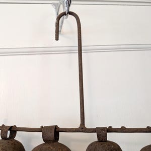 K&ouml;nnte beinhalten: Rustikaler Metall-Glocken-Windspiel mit einem gebogenen oberen Haken zum Aufh&auml;ngen. Das Windspiel hat eine horizontale Stange mit mehreren befestigten Glocken. Das Metall hat eine verwitterte, braune Oberfl&auml;che, die eine antike oder Vintage-&Auml;sthetik suggeriert.