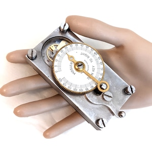 May include: A silver and brass combination lock with a white dial that reads "IN KEY TO RIGHT" and "MOSLER SWISS MADE".