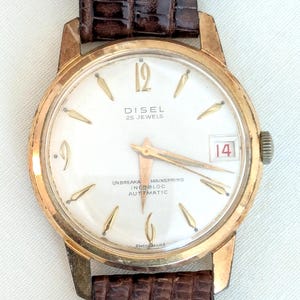 May include: A gold-tone wristwatch with a white face and black leather band. The watch face has gold-tone hands and markers, and the text "DISEL 25 JEWELS" and "UNBREAKABLE MAINSTRING INCABLOC AUTOMATIC" is printed on the face. The date window shows the number "14".