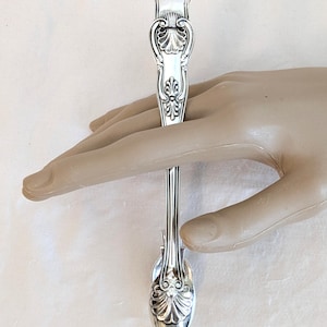 Op de afbeelding: Een zilveren lepel met sierlijke bloemen- en krulwerkdetails. De lepelsteel heeft een decoratief ontwerp en de kom heeft een afgeronde, ovale vorm. De lepel wordt tegen een neutrale achtergrond gehouden, ongeveer 15 cm lang.