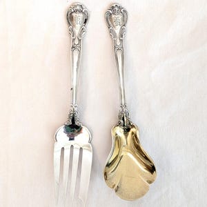 Op de afbeelding: Een zilveren serveervork en -lepelset. De handvatten hebben een sierlijk ontwerp met een initiaal. De vork heeft drie tanden en de lepel heeft een goudkleurige kom. De set wordt gepresenteerd op een wit oppervlak.