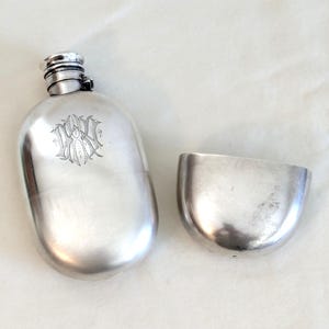 Puede incluir: Un frasco de cadera de plata con una tapa con bisagras y una taza. El frasco tiene un monograma grabado.
