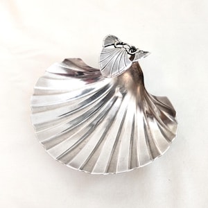 Puede incluir: Un plato plateado en forma de concha con un detalle decorativo de mariposa detallado. El plato tiene una textura acanalada y un diseño curvo y abierto. La mariposa está unida al borde superior del plato.