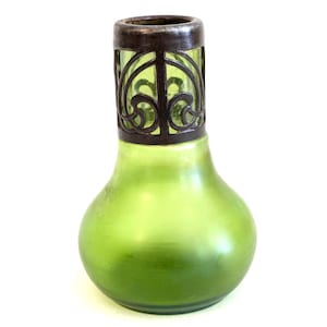 Puede incluir: Una botella de vidrio verde con base bulbosa y tapa de filigrana de metal oscuro. La botella tiene un acabado liso y mate y una forma redondeada. La parte superior de metal presenta un diseño calado ornamental.