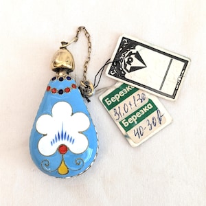 May include: A vintage, teardrop-shaped perfume bottle with a light blue enamel finish and a white floral design. The bottle has a gold-colored top and a chain. Attached tags display text in a foreign language.