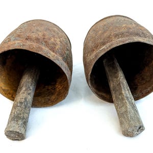 K&ouml;nnte beinhalten: Zwei antike Kuhglocken mit einem rustikalen, verwitterten Aussehen. Die Glocken bestehen aus gealtertem Metall mit einer rostbraunen Patina. Jede Glocke hat einen Holzgriff, der sich von der &Ouml;ffnung erstreckt und eine klassische, Vintage-&Auml;sthetik erzeugt.