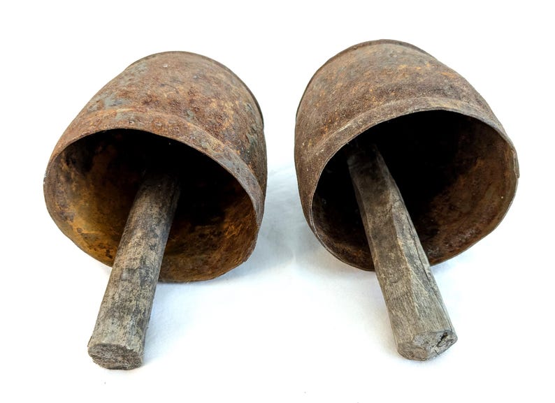 K&ouml;nnte beinhalten: Zwei antike Kuhglocken mit einem rustikalen, verwitterten Aussehen. Die Glocken bestehen aus gealtertem Metall mit einer rostbraunen Patina. Jede Glocke hat einen Holzgriff, der sich von der &Ouml;ffnung erstreckt und eine klassische, Vintage-&Auml;sthetik erzeugt.