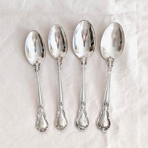 Puede incluir: Cuatro cucharas de plata con mangos ornamentados y cuencos ovalados dispuestas sobre una superficie blanca. Las cucharas tienen un acabado pulido y reflectante, y los mangos presentan diseños detallados de flores y volutas. Probablemente se trate de cubertería antigua o vintage.