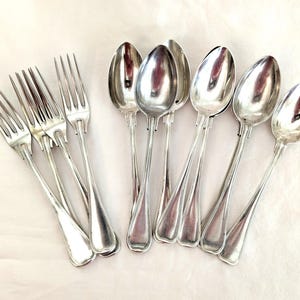 May include: A collection of polished silver forks and spoons. The forks have four tines, and the spoons have oval bowls. The silverware is arranged on a white surface, reflecting light.