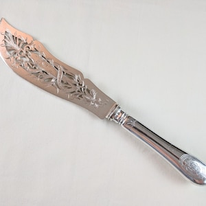 May include: A silver butter knife with a curved blade featuring intricate floral and scrollwork cutouts. The handle is smooth and rounded, with a decorative emblem at the end. The knife is designed for spreading butter or soft cheeses.