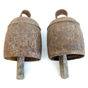 K&ouml;nnte beinhalten: Zwei antike Kuhglocken aus Metall mit verwittertem, rostigem Aussehen. Jede Glocke hat einen Schlaufengriff und einen Holzkl&ouml;ppel. Die Glocken sind in einem rustikalen Braunton gehalten und liegen vor einem wei&szlig;en Hintergrund.