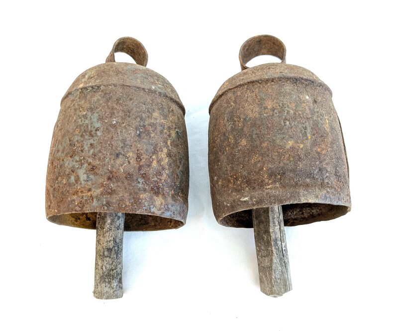 K&ouml;nnte beinhalten: Zwei antike Kuhglocken aus Metall mit verwittertem, rostigem Aussehen. Jede Glocke hat einen Schlaufengriff und einen Holzkl&ouml;ppel. Die Glocken sind in einem rustikalen Braunton gehalten und liegen vor einem wei&szlig;en Hintergrund.