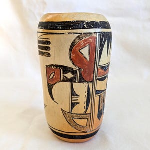 May include: A cylindrical ceramic vessel with a cream-colored base featuring hand-painted geometric and abstract motifs in black and terracotta red. The design includes bold horizontal bands at the top and bottom rim with intricate linear patterns.