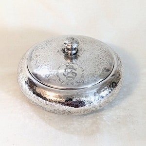 May include: A silver, hammered, round, lidded dish with a monogrammed inscription. The inscription is a circle with a rope design and the letters "BS" inside.