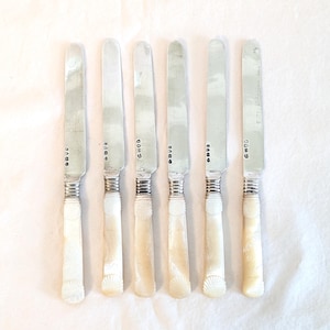 May include: A set of six antique silver knives with mother-of-pearl handles. The blades are silver, and the handles are creamy white with a shell design. The knives are arranged in a row on a white surface.