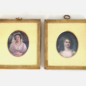 May include: Two framed oval portraits of women in ornate gold frames with a Greek key design. The portraits are painted in a realistic style and feature women with long hair and elaborate clothing.