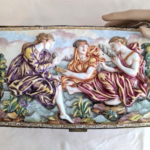 May include: Rectangular ceramic box with a detailed relief of three figures in classical attire. The figures are in shades of yellow, purple, and red, set against a backdrop of a blue sky and foliage. The box has a gold-colored trim.
