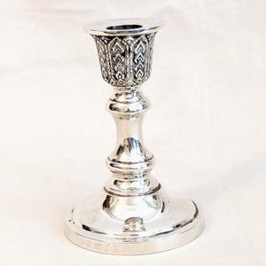 May include: A silver candlestick with a detailed, patterned cup and a round base. The candlestick has a classic design, with a smooth, reflective surface. The cup is decorated with an intricate, repeating design. The base is wide and stable.