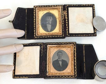 May include: Two antique black leather photo cases, each with a small portrait in an oval frame. The frames have gold trim and are set against a cream-colored background. The cases are open, revealing the portraits and a blank space.