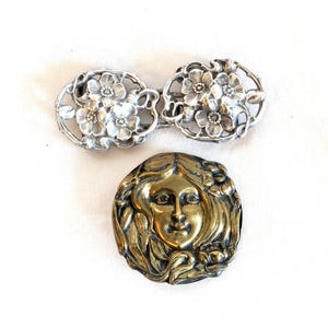 May include: A silver-toned metal two-piece belt buckle featuring embossed floral motifs and a circular brass-toned metal brooch depicting a stylized human face surrounded by flowing hair and floral elements in a relief design.
