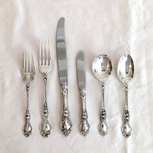 Puede incluir: Un juego de seis cubiertos antiguos de color plateado, que incluye tenedores, cuchillos y cucharas. Los mangos presentan un diseño floral ornamentado. La cubertería está dispuesta sobre una superficie blanca, mostrando la calidad reflectante del metal.