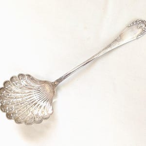 Puede incluir: Cuchara de servir plateada con un cuenco en forma de concha y un mango largo y delgado. El cuenco presenta un patrón detallado en forma de abanico y el extremo del mango es ornamentado. Una pieza vintage.