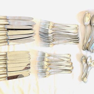 May include: A collection of silver-colored flatware, including knives, forks, and spoons, arranged on a white surface. The knives have a rectangular handle design. The forks and spoons have a classic, elegant shape. The set includes dinner and smaller spoons.