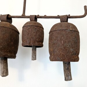 K&ouml;nnte beinhalten: Drei rostige Metallglocken mit Holzkl&ouml;ppeln, die an einer Schmiedeeisenstange h&auml;ngen. Die Glocken sind tiefbraun und weisen Gebrauchsspuren auf. Das Design deutet auf eine Vintage- oder Bauernhaus&auml;sthetik hin.