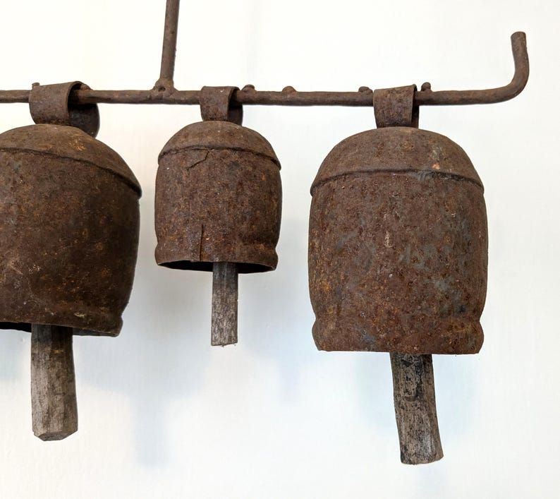 K&ouml;nnte beinhalten: Drei rostige Metallglocken mit Holzkl&ouml;ppeln, die an einer Schmiedeeisenstange h&auml;ngen. Die Glocken sind tiefbraun und weisen Gebrauchsspuren auf. Das Design deutet auf eine Vintage- oder Bauernhaus&auml;sthetik hin.