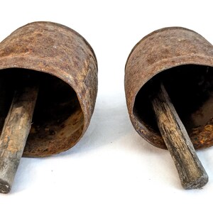 K&ouml;nnte beinhalten: Zwei antike, rostige Kuhglocken aus Metall mit Holzgriffen. Die Glocken haben eine zylindrische Form mit einer ausgestellten &Ouml;ffnung und sind vor einem wei&szlig;en Hintergrund platziert. Die Holzgriffe sind zylindrisch.