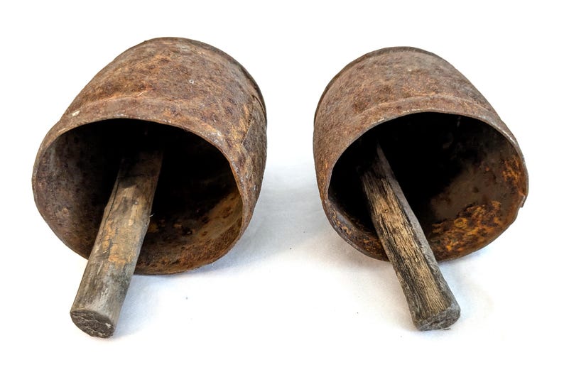 K&ouml;nnte beinhalten: Zwei antike, rostige Kuhglocken aus Metall mit Holzgriffen. Die Glocken haben eine zylindrische Form mit einer ausgestellten &Ouml;ffnung und sind vor einem wei&szlig;en Hintergrund platziert. Die Holzgriffe sind zylindrisch.